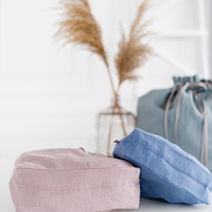 May include: Two linen tote bags, one in a light pink color and the other in a light blue color. The bags are sitting on a white surface.