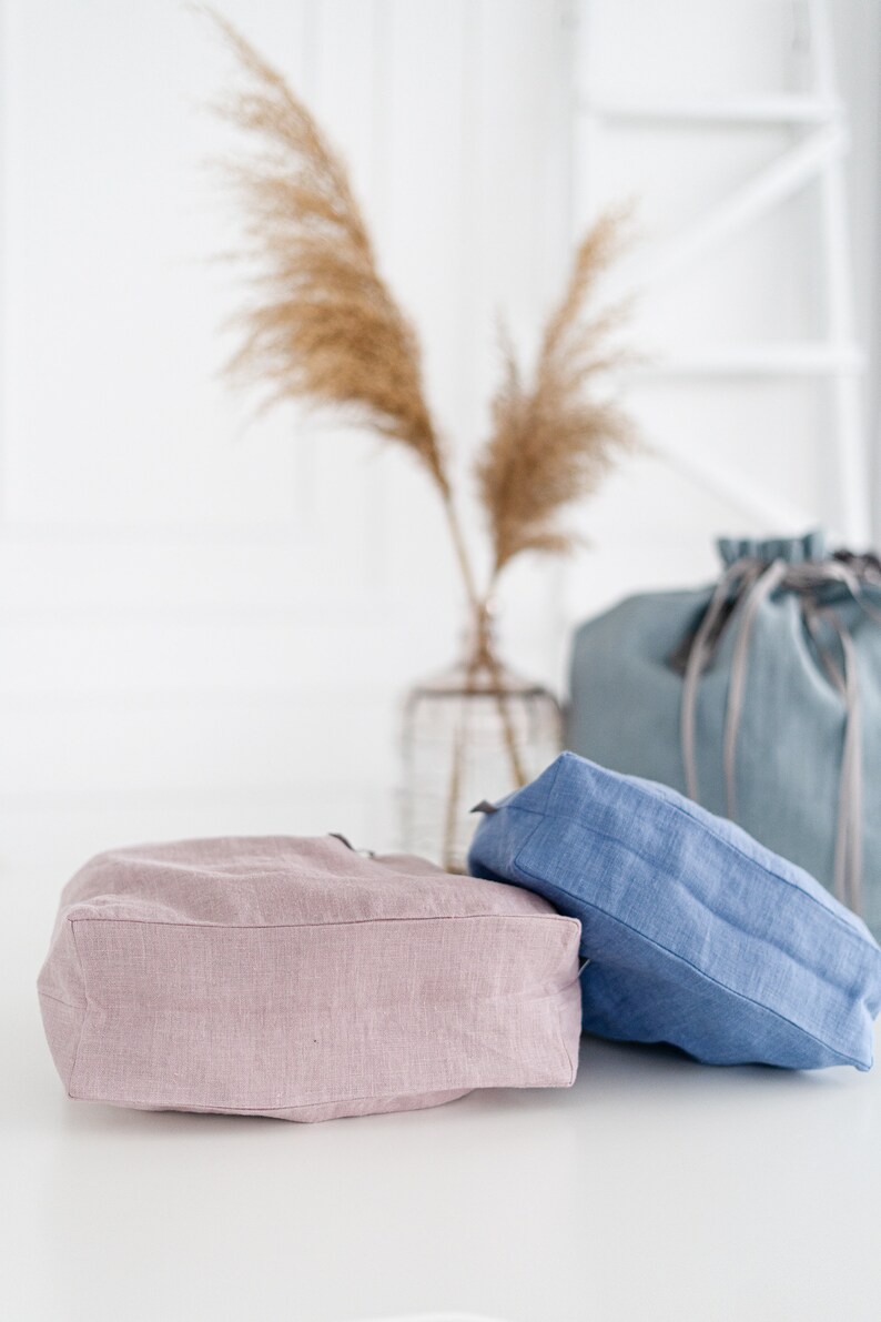 May include: Two linen tote bags, one in a light pink color and the other in a light blue color. The bags are sitting on a white surface.