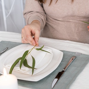 May include: A white napkin with a sprig of green leaves placed on top of a white plate. The plate is on a light blue placemat. A silver knife is on the placemat. A white candle is in the foreground.