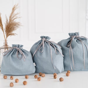 May include: Three blue linen drawstring bags with gray ribbon ties. The bags are arranged in a row on a white surface with walnuts scattered around them.