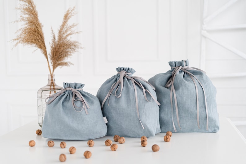 May include: Three blue linen drawstring bags with gray ribbon ties. The bags are arranged in a row on a white surface with walnuts scattered around them.
