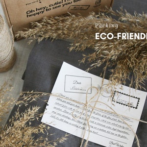 May include: A gray linen fabric package tied with twine and a handwritten note that says "Dear Anna" and "Best Wishes". The package is surrounded by dried grass and the text "Packing ECO-FRIENDLY" is visible in the background.