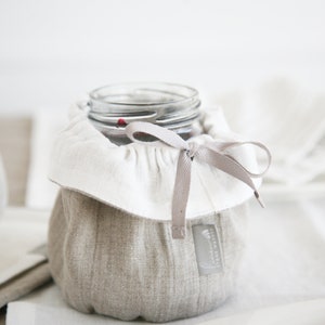 Puede incluir: Un tarro de vidrio con una cubierta de lino y una cinta gris atada alrededor de la parte superior. El tarro está sentado sobre una superficie blanca.