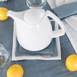 Può includere: Una teiera bianca su un sottopentola di lino blu con un bordo di lino bianco. Due limoni gialli sono sul tavolo.