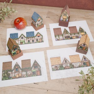 Four folded paper models of country cottages rest on a light wood table next to their unfolded paper templates. An apple in the top left corner gives a sense of scale for the miniature houses. The composition is set against a reddish wall and decorated with green plant branches entering the frame from the sides.