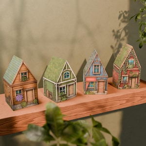 May include: Four detailed miniature paper houses are displayed on a wooden shelf. The houses are painted in various colours, including brown, green, and blue, with green roofs and small windows. Each house has a unique design, creating a charming village scene.