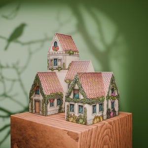 Puede incluir: Una colección de casas en miniatura con tejados de tejas rojas y fachadas detalladas, adornadas con enredaderas y flores. Las casas están dispuestas sobre un bloque de madera, creando una encantadora escena de pueblo. El fondo es verde.