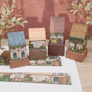 Four paper models of rustic cottages arranged in a line. The first on the left has large, rounded windows. Next to the second, gardening tools, a green backpack, and a hat rest on a wooden wheelbarrow. The third cottage features stacked hay bales with pumpkins and a watermelon, alongside bundles of brushwood. The fourth cottage on the right has sacks on wooden platforms and a pergola with ripe grapes.