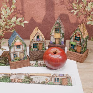 This image shows four folded models of paper huts, standing on wooden stands, having slightly different heights. The huts are visible from the front, and in front of them lies a red apple, placed there as a comparison of the scale. The houses have different heights, but they are all slightly larger than the apple, especially when it comes to height.