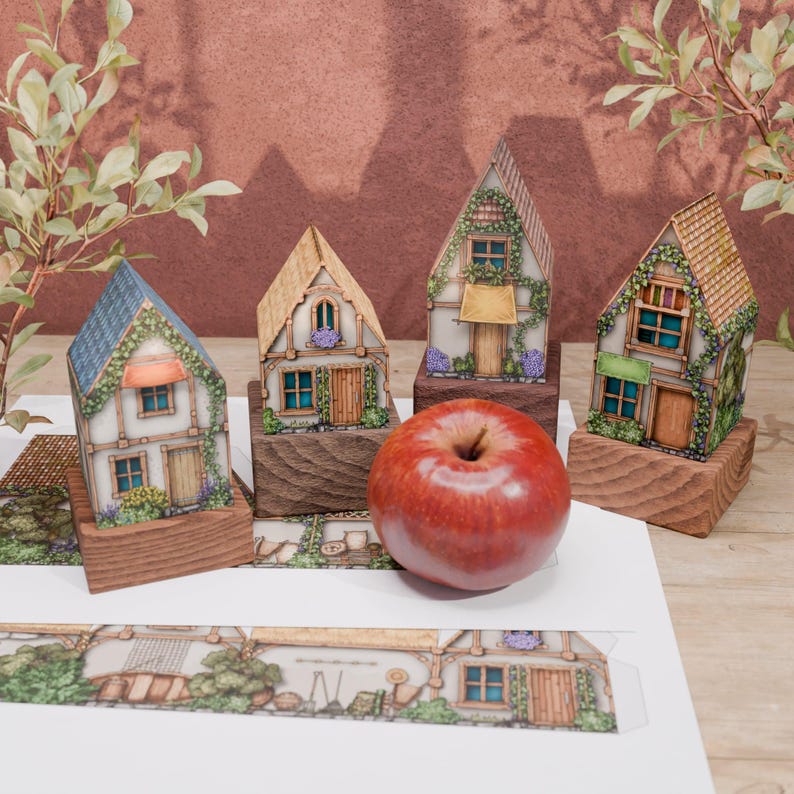 This image shows four folded models of paper huts, standing on wooden stands, having slightly different heights. The huts are visible from the front, and in front of them lies a red apple, placed there as a comparison of the scale. The houses have different heights, but they are all slightly larger than the apple, especially when it comes to height.