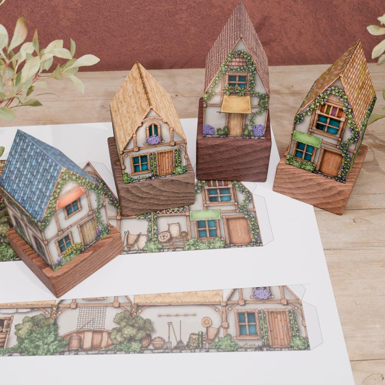 Four paper models of country cottages are shown from an angle. In the foreground are two unfolded paper templates, revealing the simple, one-piece design for easy assembly. The models feature a traditional illustration style with detailed textures of rustic life, including gardening tools, flowers, a wheelbarrow, and a small boat.