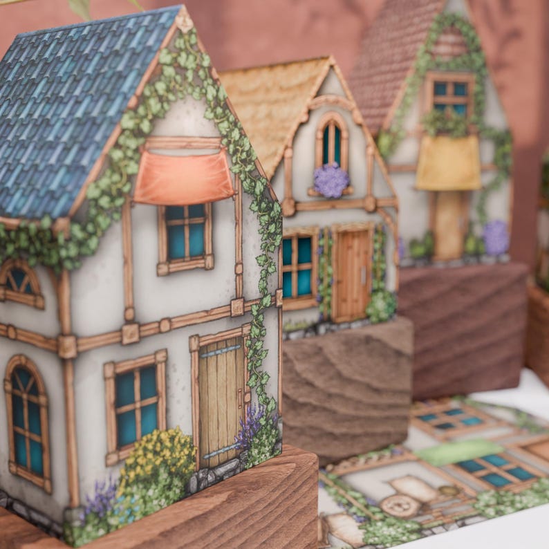 A close-up of three paper cottages arranged one behind the other, with a soft focus on the one in front. The closest cottage has a blue-tiled roof, an orange awning, and its walls are covered with climbing vines and flowers. In the background, two other cottages with thatched and tiled roofs are visible, along with a burgundy wall showing shadows of a window and plants from outside the frame. The models show a high level of hand-drawn detail.