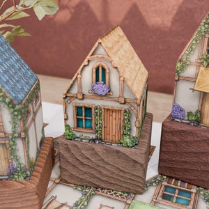 A close-up on a paper model of a cottage with a thatched roof. The ground floor has a wooden door and a four-part window. The upper floor has a rounded window with a dense cluster of pink flowers on its sill. Grape vines grow on two pergolas on either side of the front door, enhancing the idyllic, rustic style of the cottage.