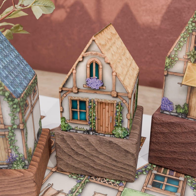 A close-up on a paper model of a cottage with a thatched roof. The ground floor has a wooden door and a four-part window. The upper floor has a rounded window with a dense cluster of pink flowers on its sill. Grape vines grow on two pergolas on either side of the front door, enhancing the idyllic, rustic style of the cottage.