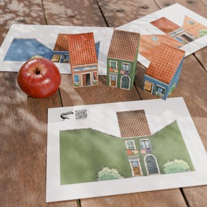 Op de afbeelding: Drie miniatuur papieren huizen met rode pannendaken en kleurrijke gevels staan op een houten oppervlak, naast een rode appel. Huis sjablonen en een logo zijn zichtbaar op de achtergrond.