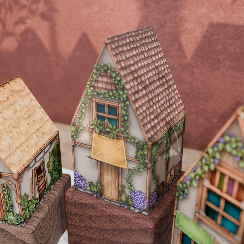 A close-up of a paper cottage model with a pointed, red-tiled roof, resting on a dark wood base. The cottage is covered in climbing vines, with purple flowers at its base and ferns on the upper window sill. A yellow awning hangs over the front door. On either side, parts of two other cottages are visible. In the background, a textured burgundy wall displays shadows from the models and off-screen foliage.