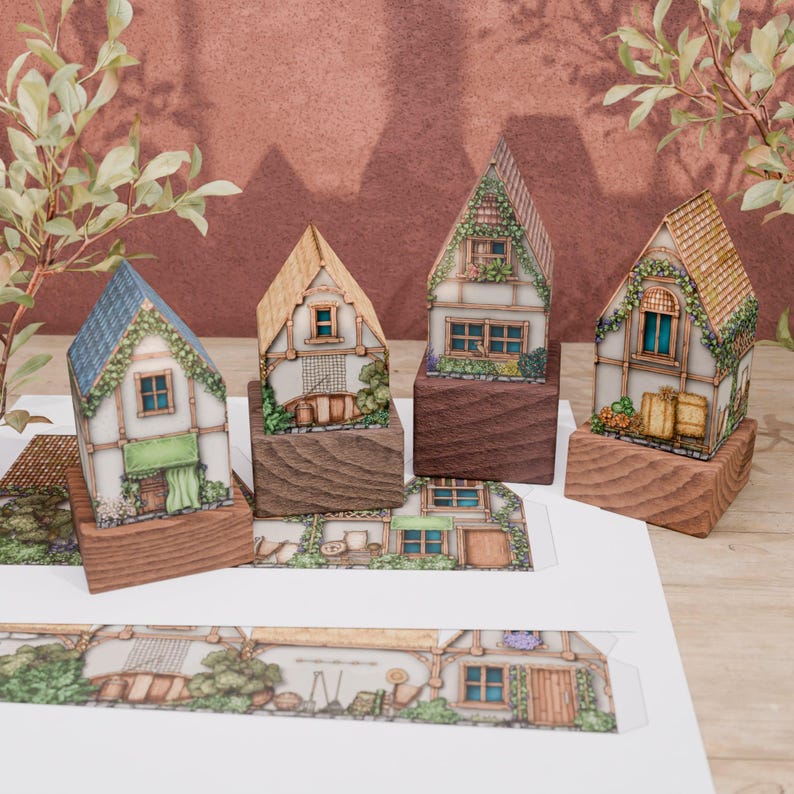 Four folded paper models of country huts on wooden stands. Rear windows with wooden frames, hay bales, pumpkins, watermelons, lettuce, and hay sheaves are visible. Flowers grow under the windows. A small boat, rod, net, and barrel lean against one hut, suggesting water-related occupations. This hut has a light thatch roof, while others have tile roofs. All huts have stone foundations.