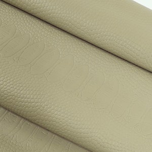 May include: Close-up of a beige faux leather fabric with a textured surface. The fabric is folded in thirds, showing the texture and colour.