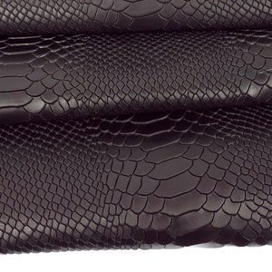 Op de afbeelding: Een close-up van een donkerbruine imitatie slangenhuid stof. De stof is in drieën gevouwen, waardoor de textuur van het materiaal zichtbaar is.