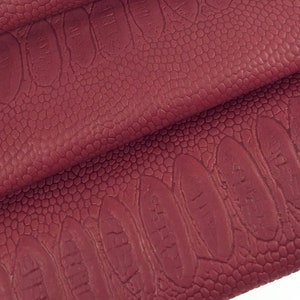 Könnte beinhalten: Burgunderfarbener Kunstlederstoff mit einem geprägten Krokodilmuster. Der Stoff ist dreifach gefaltet und zeigt die Textur des Materials.