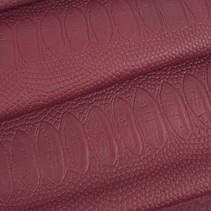 May include: Burgundy faux leather fabric with an embossed crocodile pattern. The fabric is folded in layers, showing the texture and colour.