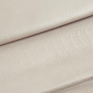May include: Close-up of folded, light taupe leather with an embossed ostrich pattern. The leather has a textured surface with small, round bumps and larger, oval indentations. The folds create shadows, highlighting the texture and colour variations.