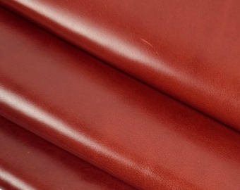 May include: Close-up of three overlapping sheets of red-brown leather. The leather has a smooth, shiny surface and appears to be folded over itself. The background is plain white, highlighting the rich color and texture of the leather.