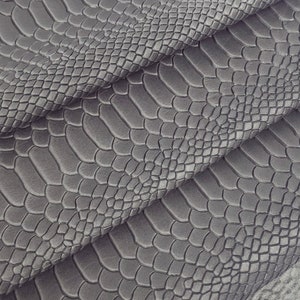 May include: Close-up of a grey snakeskin-embossed leather fabric. The fabric is folded in layers, showing the texture and pattern.