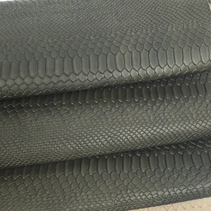 Op de afbeelding: Close-up van donkergrijze imitatie slangenleer vellen. Het leer heeft een textuur, schubbenachtig patroon. De vellen zijn gelaagd, waardoor een lichter beige leer eronder zichtbaar is. De afbeelding is tegen een witte achtergrond.