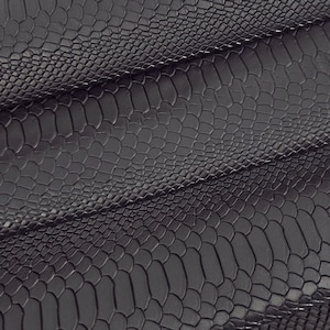 Può includere: Primo piano di pelle nera con motivo a pelle di serpente. La pelle è piegata, rivelando la consistenza e la lucentezza del materiale. La superficie presenta un disegno a rilievo a forma di scaglie. Lo sfondo è bianco.