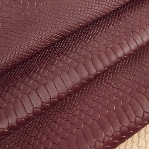 Könnte beinhalten: Nahaufnahme eines burgunderfarbenen Kunstleders mit Schlangenhautmuster. Der Stoff hat eine strukturierte, geprägte Oberfläche, die an Schlangenschuppen erinnert. Der Stoff ist dreifach gefaltet und zeigt die Unterseite des Stoffes, die hellbeige ist.