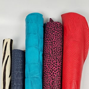 Könnte beinhalten: Eine Sammlung gerollter Lederhäute in verschiedenen Farben und Mustern. Enthält Zebra-Print, marineblaues Krokodil, türkisfarbenes Krokodil, pinkfarbenen Leoparden-Print und rotes Straußenleder. Die Häute sind vertikal vor weißem Hintergrund angeordnet.
