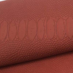 May include: Close-up of a textured, reddish-brown leather with a subtle embossed pattern. The leather is folded in thirds, revealing the texture and colour.