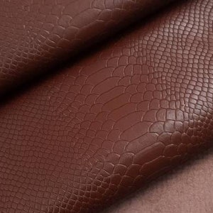 May include: A close-up of a brown faux leather fabric with a crocodile embossed pattern. The fabric is folded over itself, revealing a brown suede backing.