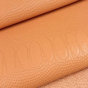 Puede incluir: Primer plano de una tela de piel sintética texturizada de color melocotón. La tela tiene un sutil patrón en relieve que se asemeja a escamas.