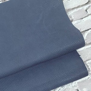 Può includere: Primo piano di due pezzi sovrapposti di pelle blu con una superficie strutturata. La pelle è di un colore blu scuro uniforme. Lo sfondo è un muro di mattoni bianchi.