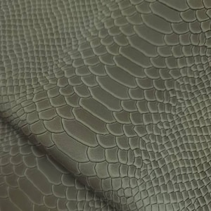 Op de afbeelding: Close-up van een vel olijfgroen imitatieleer met een reptielschubbenpatroon. Het materiaal heeft een getextureerd oppervlak met een subtiele glans en is in de linkerbenedenhoek gevouwen.