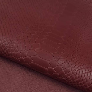 Puede incluir: Primer plano de una tela de imitación de cuero burdeos con un patrón en relieve de cocodrilo. La tela está doblada para revelar un respaldo de ante burdeos suave.