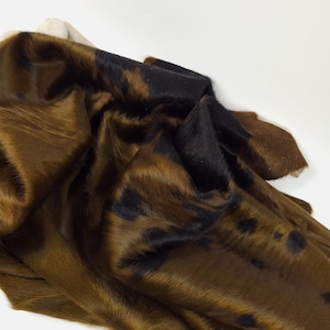 May include: A folded piece of cowhide in shades of brown, black, and tan. The cowhide has a soft, furry texture and a natural pattern. The colours are rich and earthy, with dark spots scattered across the surface.