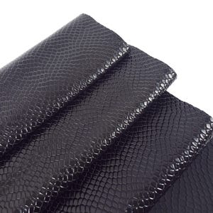 May include: Close-up of black faux snakeskin fabric. The material has a textured, scale-like pattern and a glossy finish. The fabric is folded over itself, revealing the texture and sheen. Ideal for fashion and craft projects.