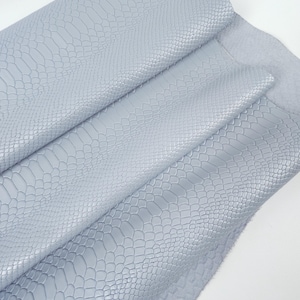 May include: Pale blue leather with a reptile-like embossed pattern. The leather is folded, showing the texture and colour variations. The material appears soft and pliable, suitable for various crafting projects.