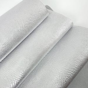 Könnte beinhalten: Silberfarbenes Kunstleder mit einem strukturierten, schuppenartigen Muster. Das Material ist gerollt und gefaltet, wodurch seine Flexibilität und sein Glanz zur Geltung kommen. Der Stoff hat ein metallisches Aussehen und reflektiert das Licht. Ideal für Bastelprojekte.