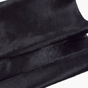 Könnte beinhalten: Nahaufnahme eines gefalteten Stücks schwarzem Leder. Das Leder hat eine weiche, strukturierte Oberfläche mit leichtem Glanz. Die Falten erzeugen Schatten und Glanzlichter, die die Tiefe und Textur des Materials hervorheben. Der Hintergrund ist weiß.