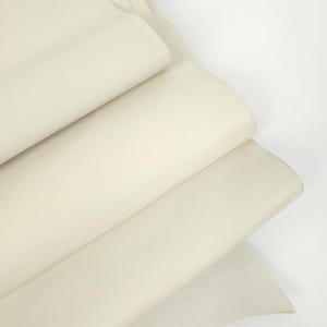 Peut inclure: Feuilles de cuir crème, empilées et partiellement enroulées, révélant une texture douce et lisse. Les bords montrent le grain naturel du matériau. La couleur neutre suggère une polyvalence pour divers projets artisanaux.