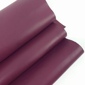 May include: A close-up of several sheets of burgundy leather. The leather has a smooth, matte finish and is folded over itself. The edges of the leather are visible, showing the thickness and texture of the material. The background is plain white.