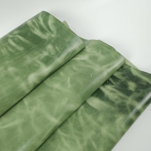 May include: Close-up of several sheets of green leather. The leather has a mottled appearance with lighter green areas, creating a textured effect. The sheets are folded and layered, showcasing the material's flexibility and color variations.