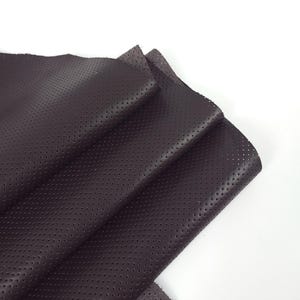 May include: Close-up of folded dark brown perforated leather. The leather has a matte finish and small, evenly spaced holes. The edges are slightly frayed, against a white background. Ideal for crafting.