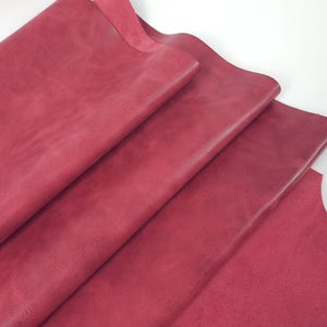 Puede incluir: Primer plano de varias hojas de cuero de color burdeos. El cuero tiene una superficie texturizada y está doblado sobre sí mismo, revelando el reverso del material en algunas zonas. El fondo es blanco liso.