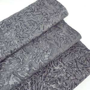 May include: Three layers of gray fabric with a textured, leaf-like pattern. The fabric has a crinkled appearance, with variations in light and dark gray tones. The material is folded, revealing the layers and the intricate design.
