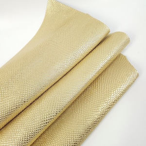 May include: Close-up of several sheets of gold-coloured faux leather. The material has a textured, scale-like pattern. The sheets are folded and layered, showcasing the material's sheen and flexibility. The background is plain white.